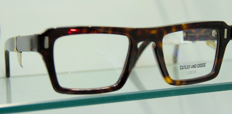 A pair of tortoiseshell, rectangular Cutler and Gross glasses on a glass shelf at the opticians