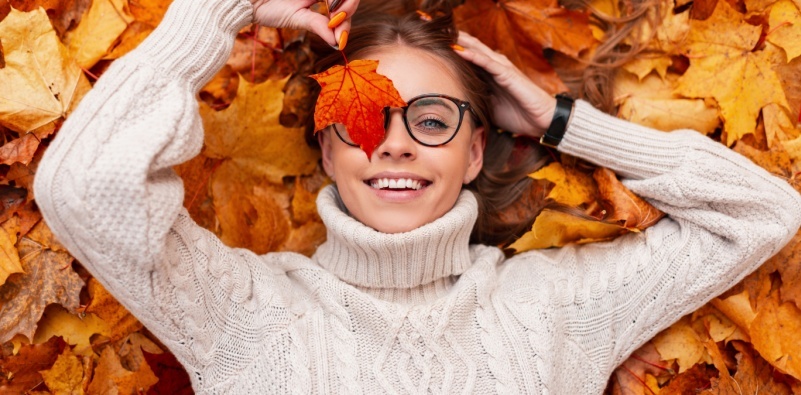 A cheerful young woman, lying on autumn leaves, in stylish sunglasses in a knitted white sweater, holding an orange lead over her eye