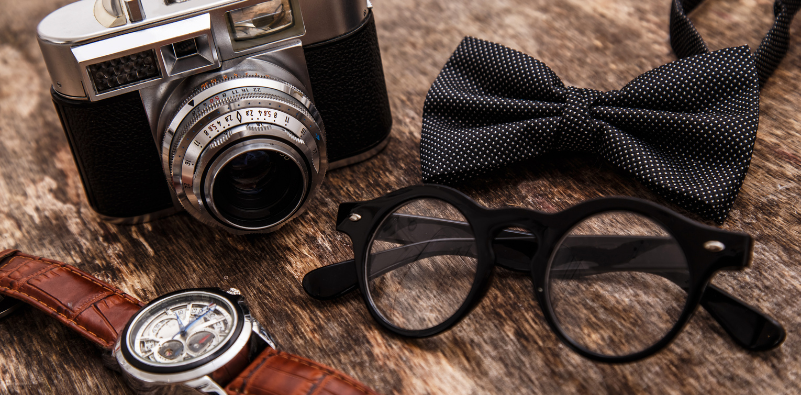 A flat lay of a pair of black, round glasses alongside a vintage cmera, watch and bow tie