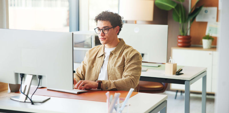 A young man wearing glasses working at a desktop computer in a bright office.